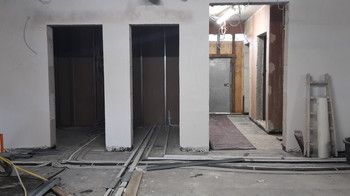 Blick auf eine Wand mit zwei leeren Türöffnungen. Die Wände sind verputzt und im Hintergrund sieht man eine unverputzte Trockenbauwand. Auf der rechten Seite sieht man noch den Flur vor dem Treppenhaus, hier sind die Wände noch unverputzt. Auf dem Boden sind Kabel verlegt.