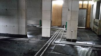 Foto vom Inneren des Rohbaus mit unverputzten Wänden. Auf dem Fußboden ist Dachpappe verlegt und einige Rohre verlaufen durch die Räume. rechts im Bild ist der Fahrstuhlschacht zu sehen, der mit einer Holzpalette abgesperrt ist.